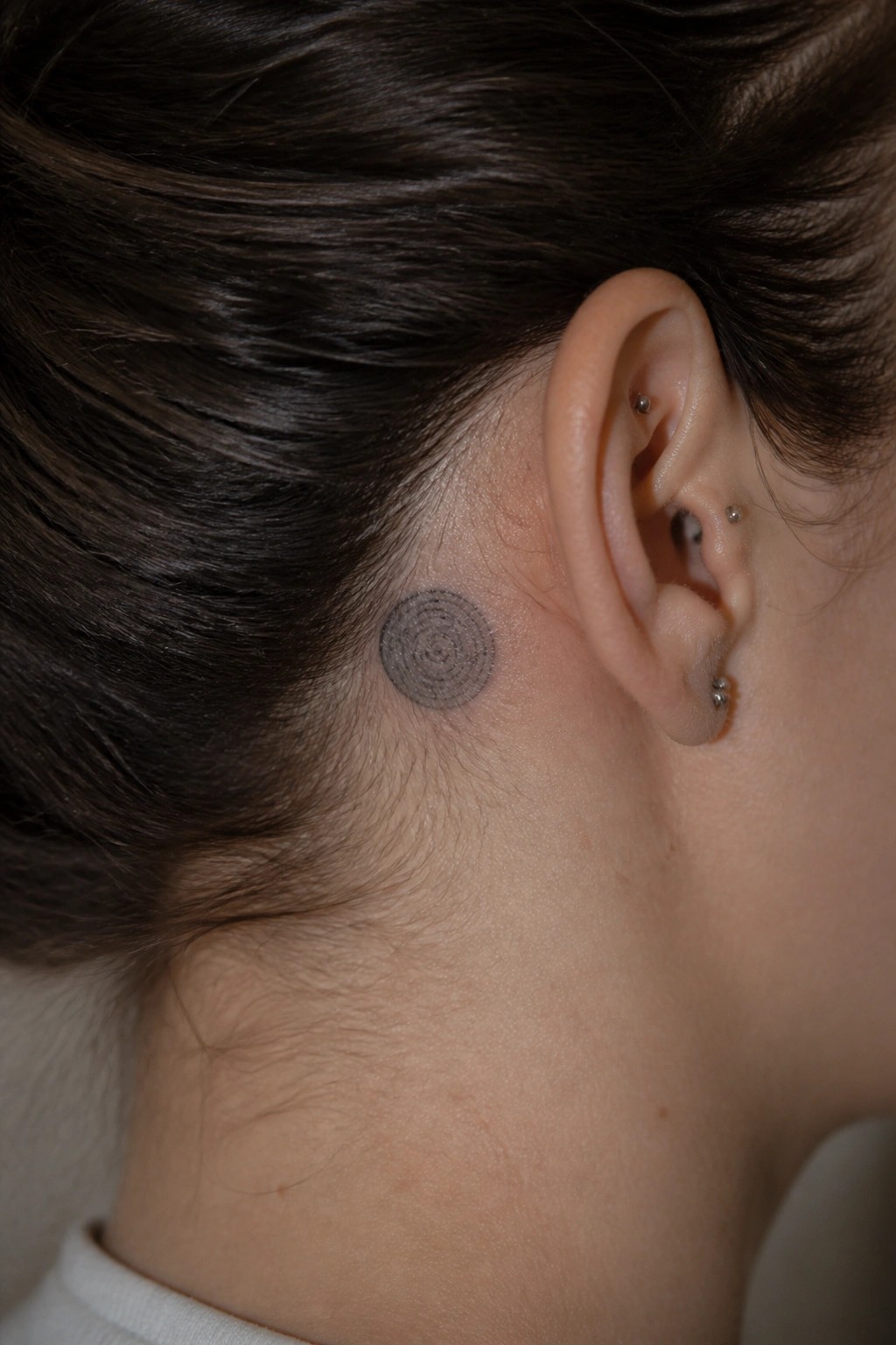 Circular black tattoo behind ear with multiple piercings on dark-haired person