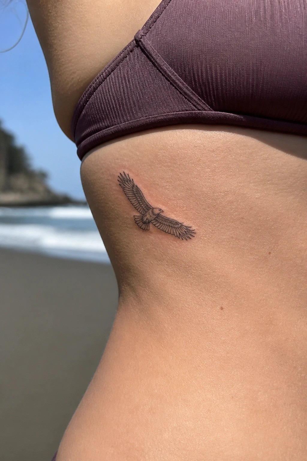 Small black eagle tattoo below purple bikini top on sunny beach skin