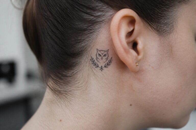 19 Meaningful Owl Tattoo Ideas for Women Inspired by Wisdom and Strength Small fine line owl tattoo with leaves placed behind the ear on dark hair.