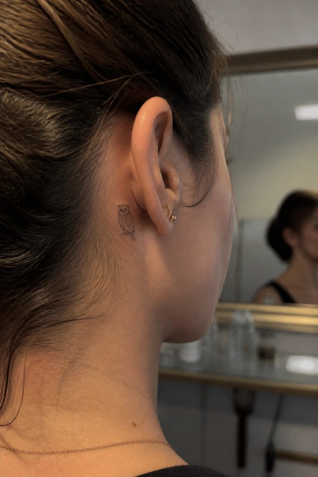 Dark-haired woman with small owl tattoo behind ear, gold earrings and necklace