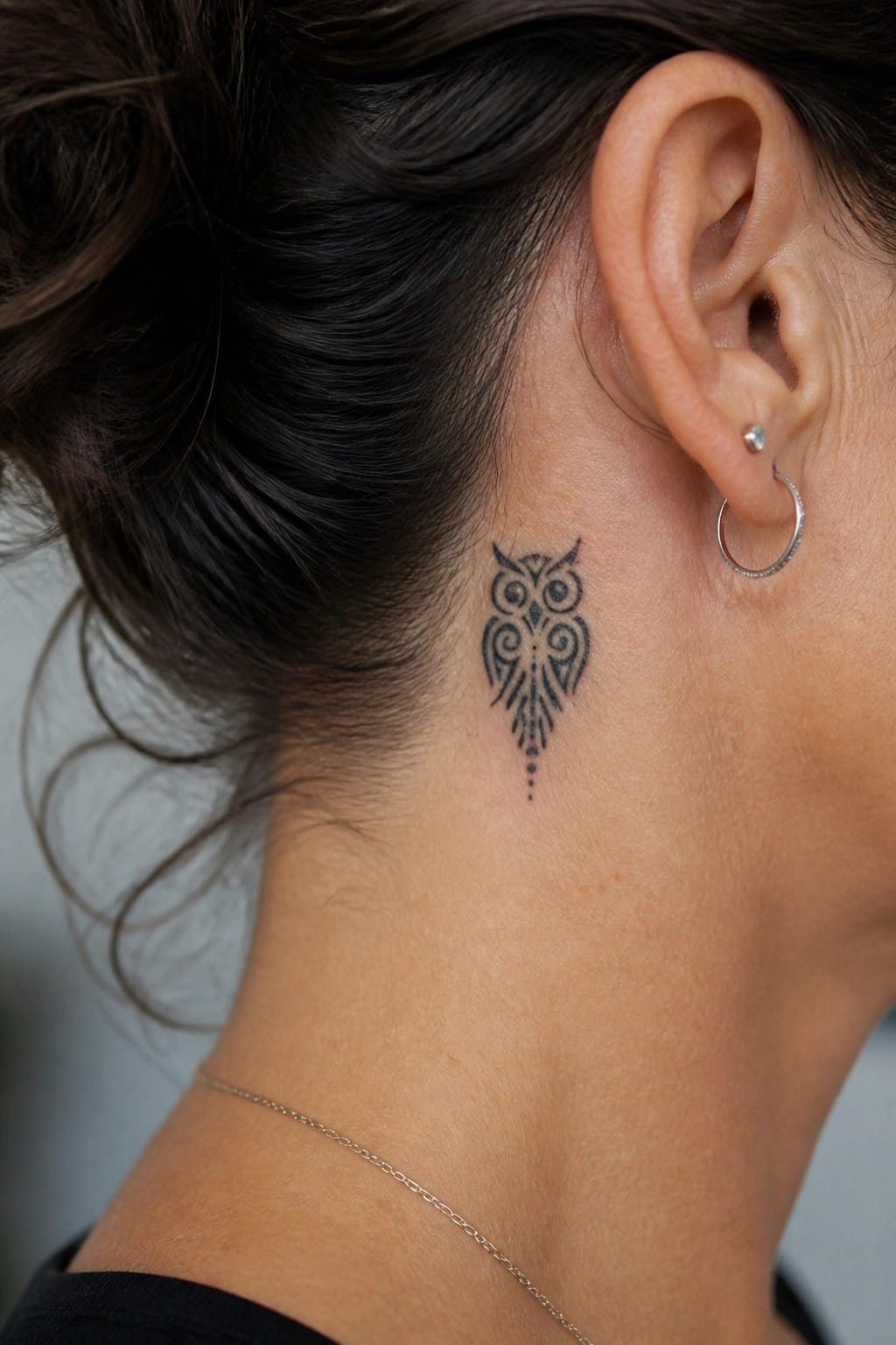 Owl tattoo on neck behind ear, dark bun, silver hoop earring, thin necklace