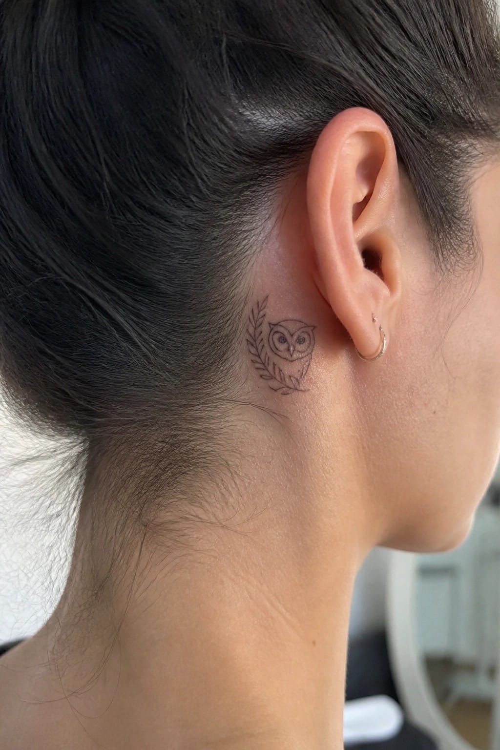 Small fine line owl tattoo with leaves placed behind the ear on dark hair.