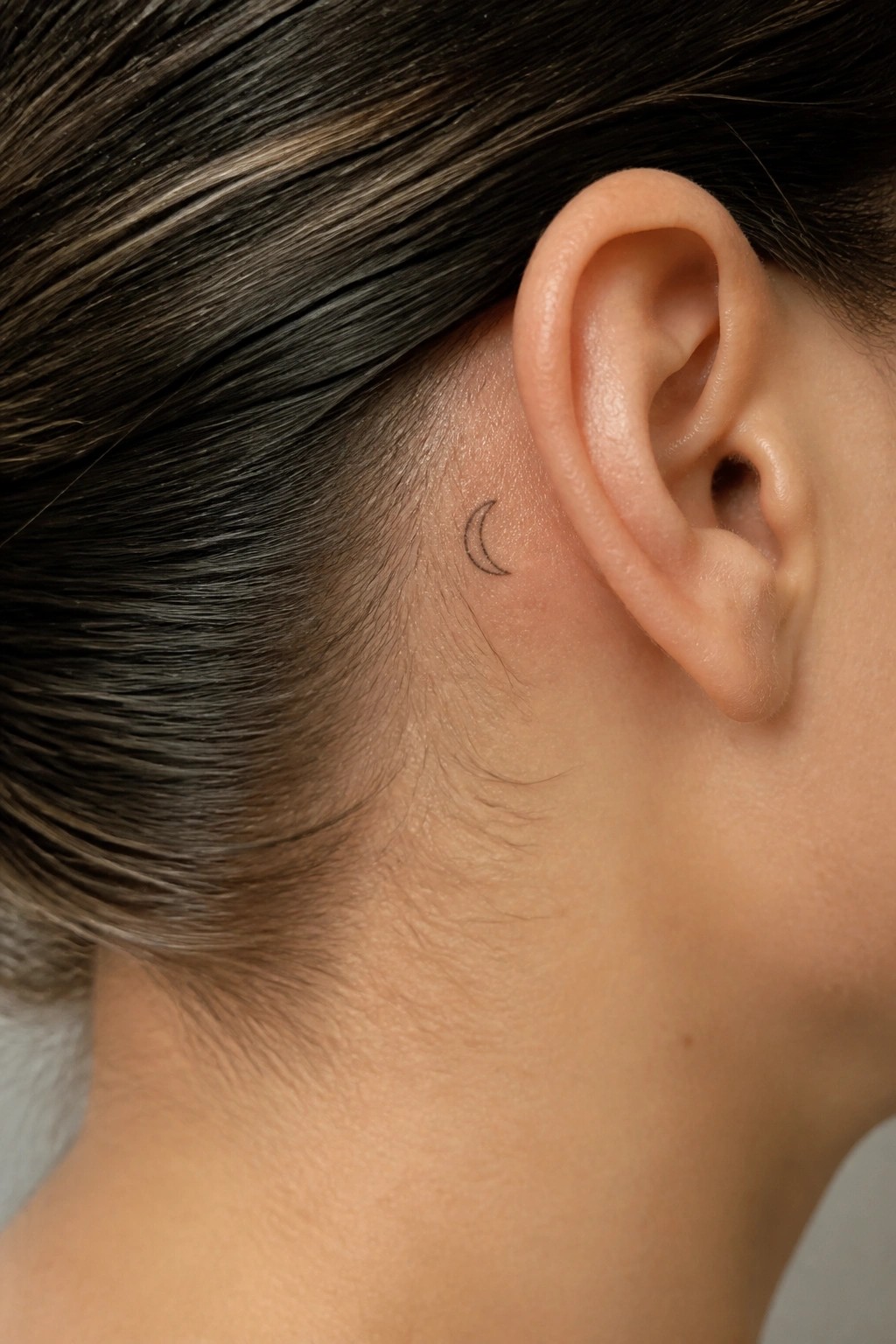 Crescent moon tattoo behind an ear on light skin with dark hair.
