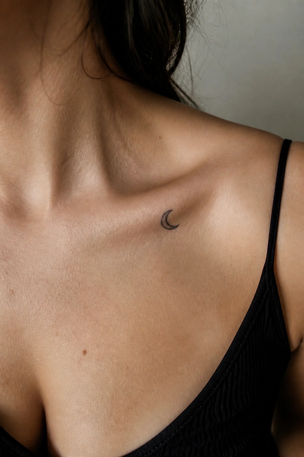 Fair-skinned shoulder with small crescent moon tattoo beside black spaghetti strap