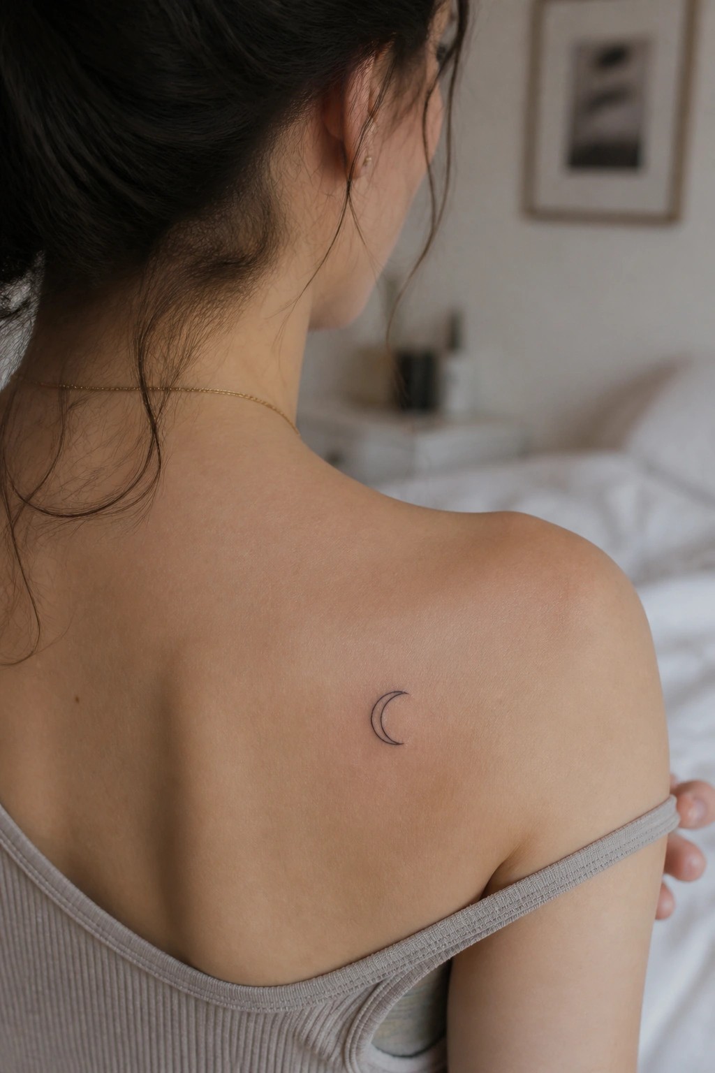 Woman's back with crescent moon tattoo on shoulder, wearing gray tank top in bedroom