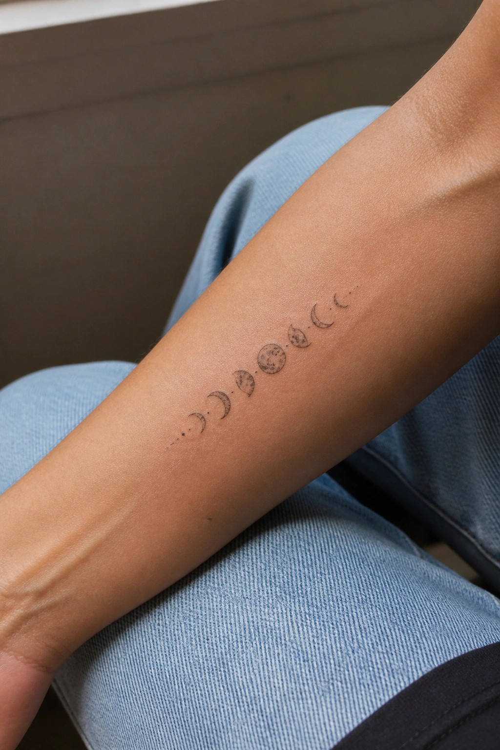 Moon phases tattoo on forearm resting atop light blue denim jeans
