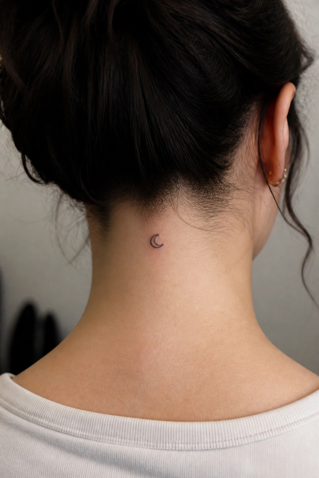 Small crescent moon tattoo on neck below dark bun, white shirt visible.