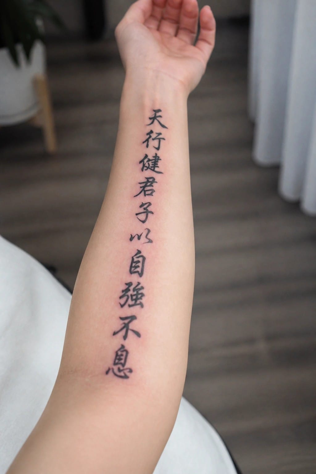 Vertical black Chinese characters tattooed along raised forearm in indoor setting.