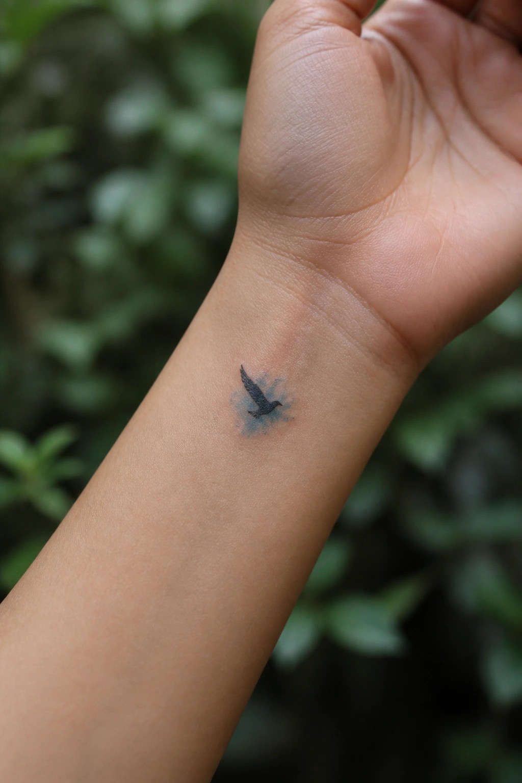 Small black flying bird tattoo on the inner wrist with light blue watercolor shading around it