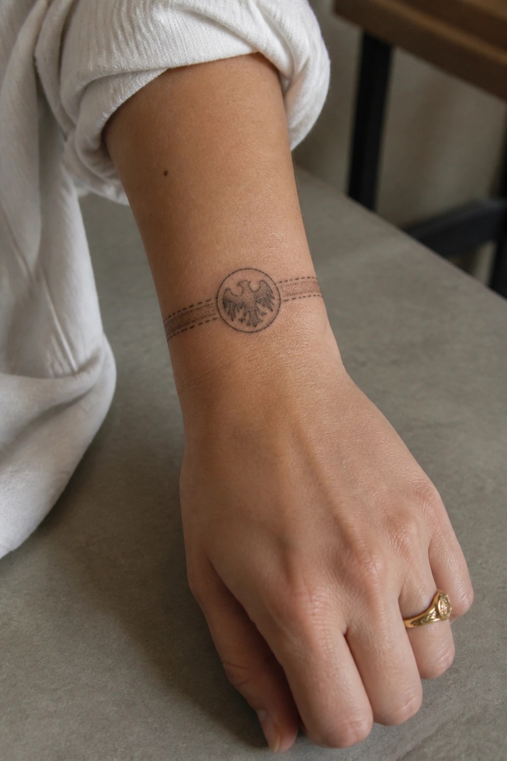 Wrist with circular double-headed eagle tattoo and dashed lines, white rolled sleeve, gold ring.