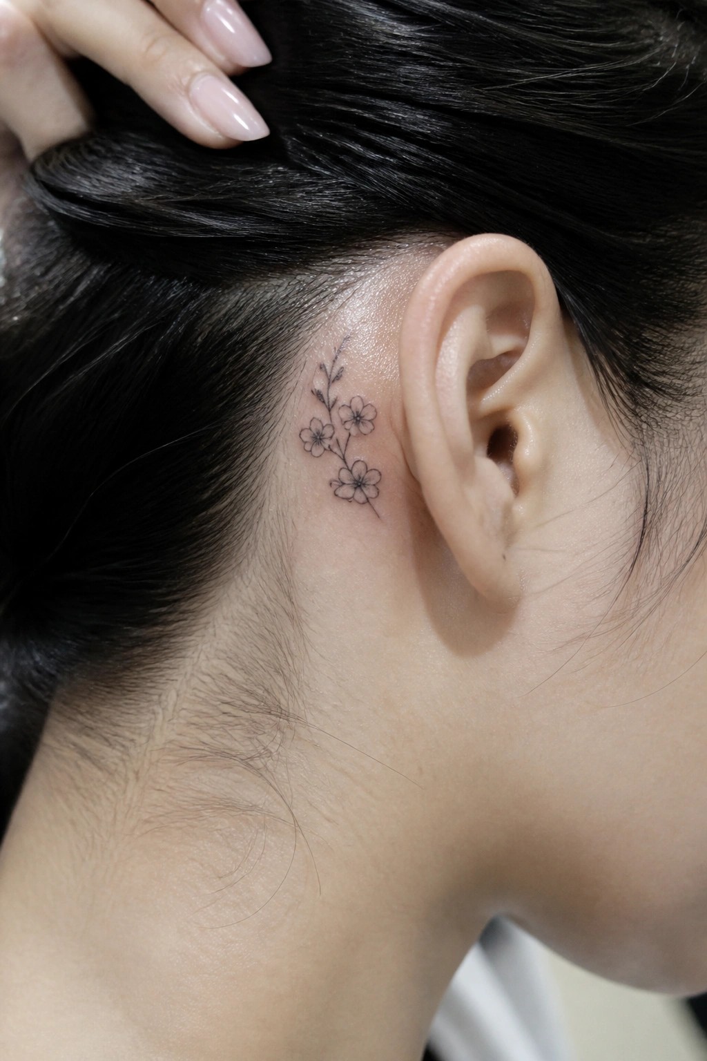 Small black floral tattoo behind ear with dark hair held by manicured hand