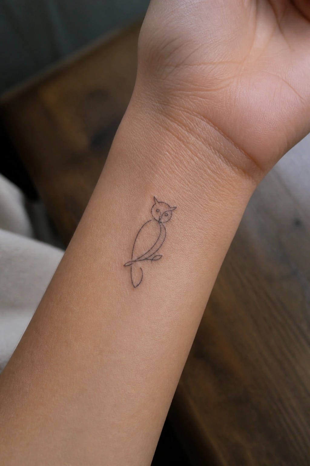Small minimalist line art owl tattoo on the inner wrist, shown perched on a simple branch.