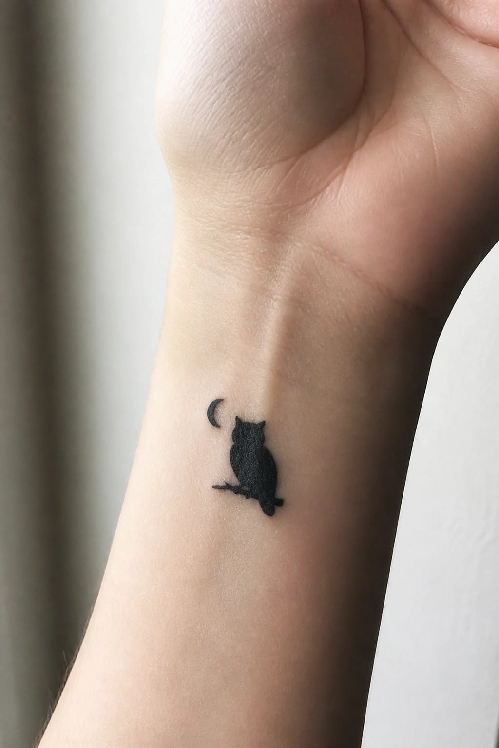 Minimalist black owl tattoo on wrist beside crescent moon on branch
