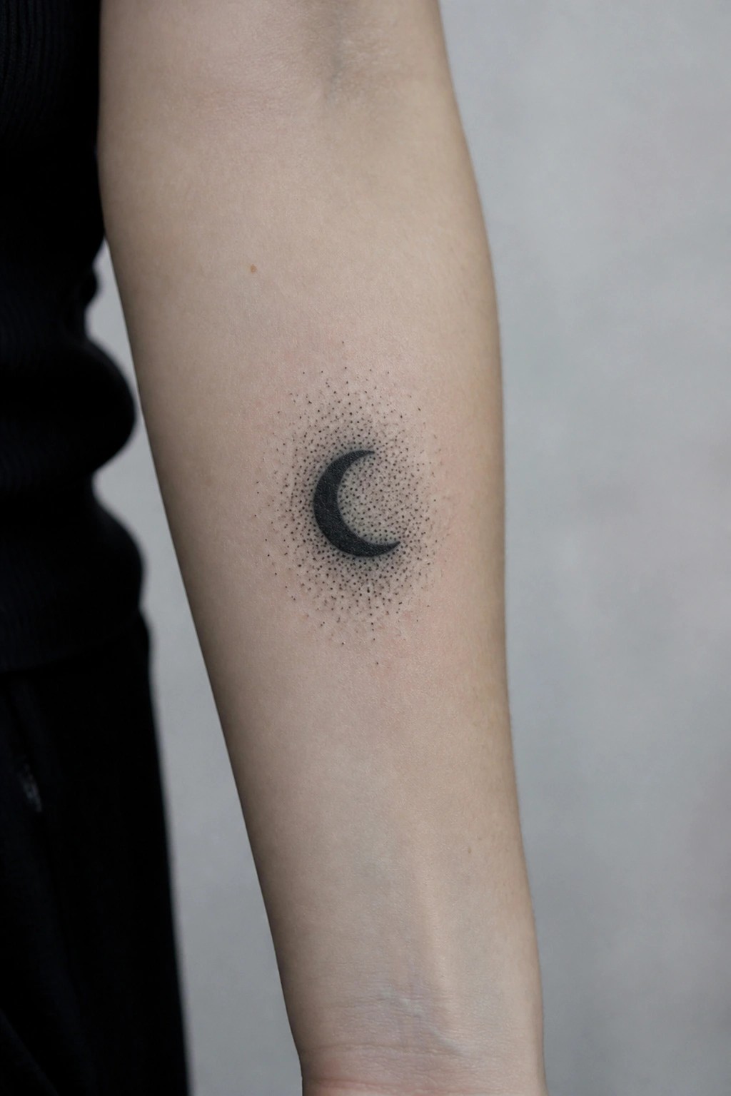 Dotted black crescent moon tattoo on a person's forearm in black clothing