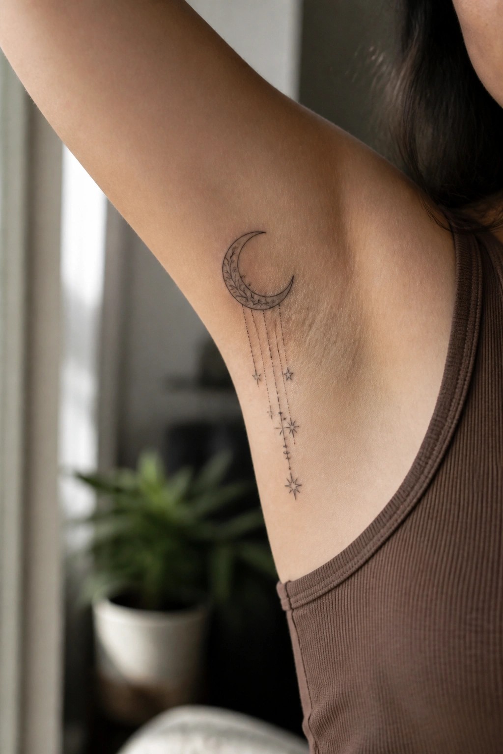 Crescent moon with leafy details and dangling stars tattoo on arm above brown tank top