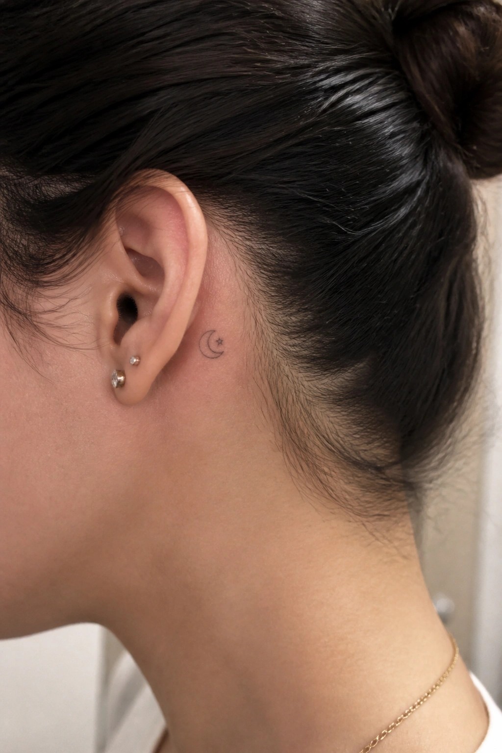Close-up of ear with crescent moon-star tattoo, two small stud earrings, gold chain.