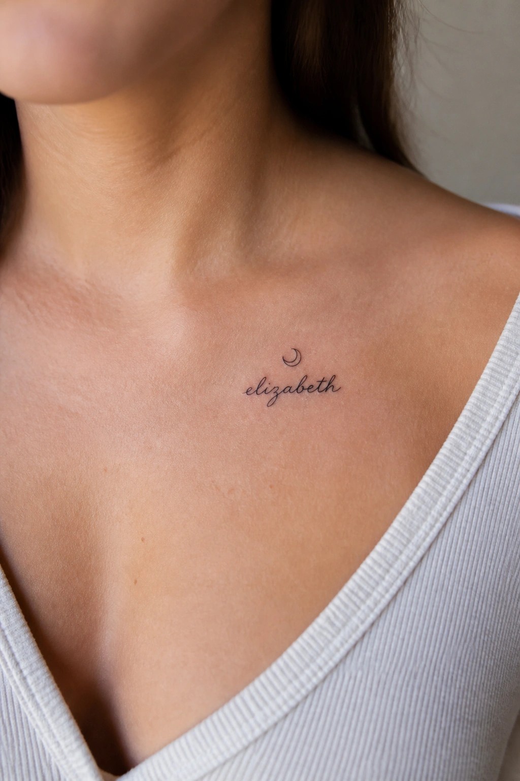 Small fine line tattoo of the name Elizabeth in cursive with a crescent moon above it placed on the upper chest.