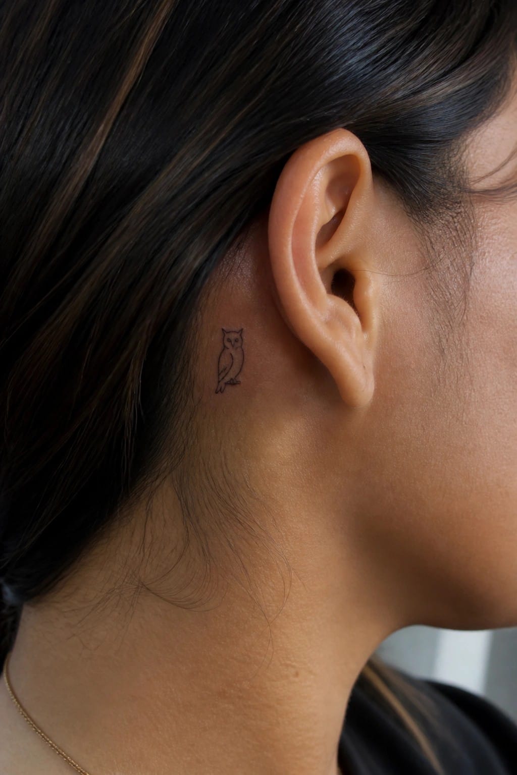 Small minimalist owl tattoo in black outline placed behind a woman's ear.