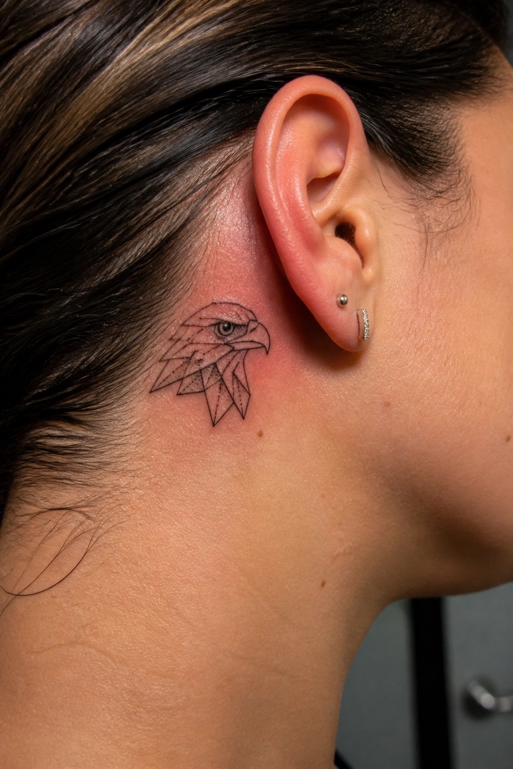 Geometric eagle tattoo behind ear on reddened skin with small earrings