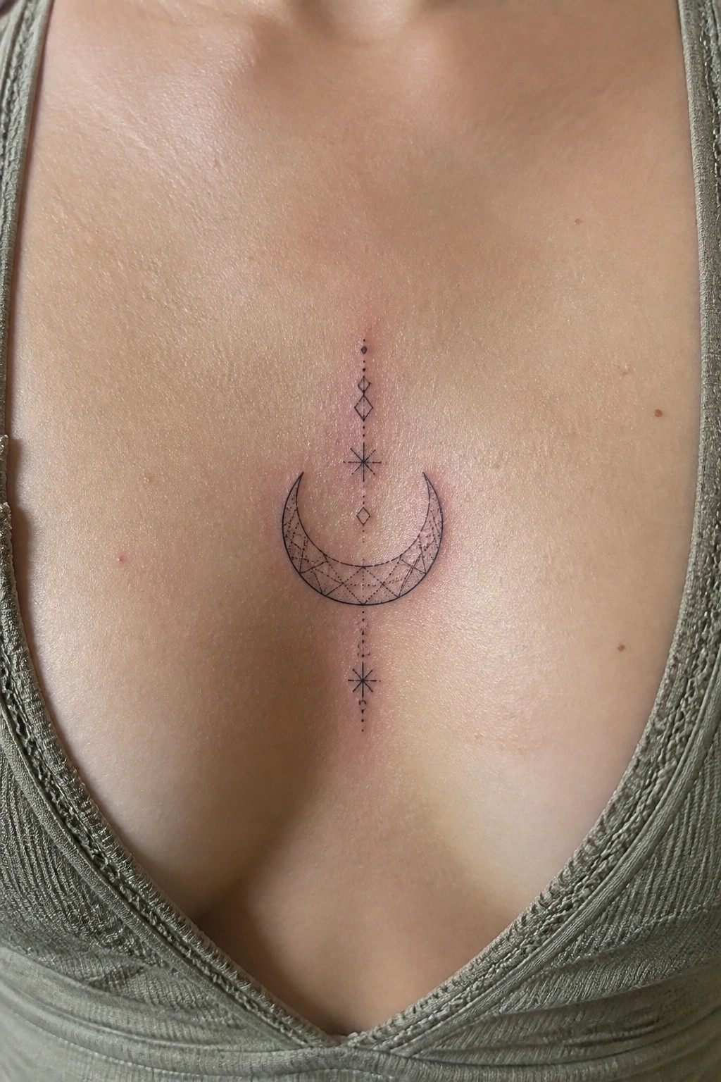 Small crescent moon tattoo with geometric patterns inside and a vertical line of dots and stars on the sternum.
