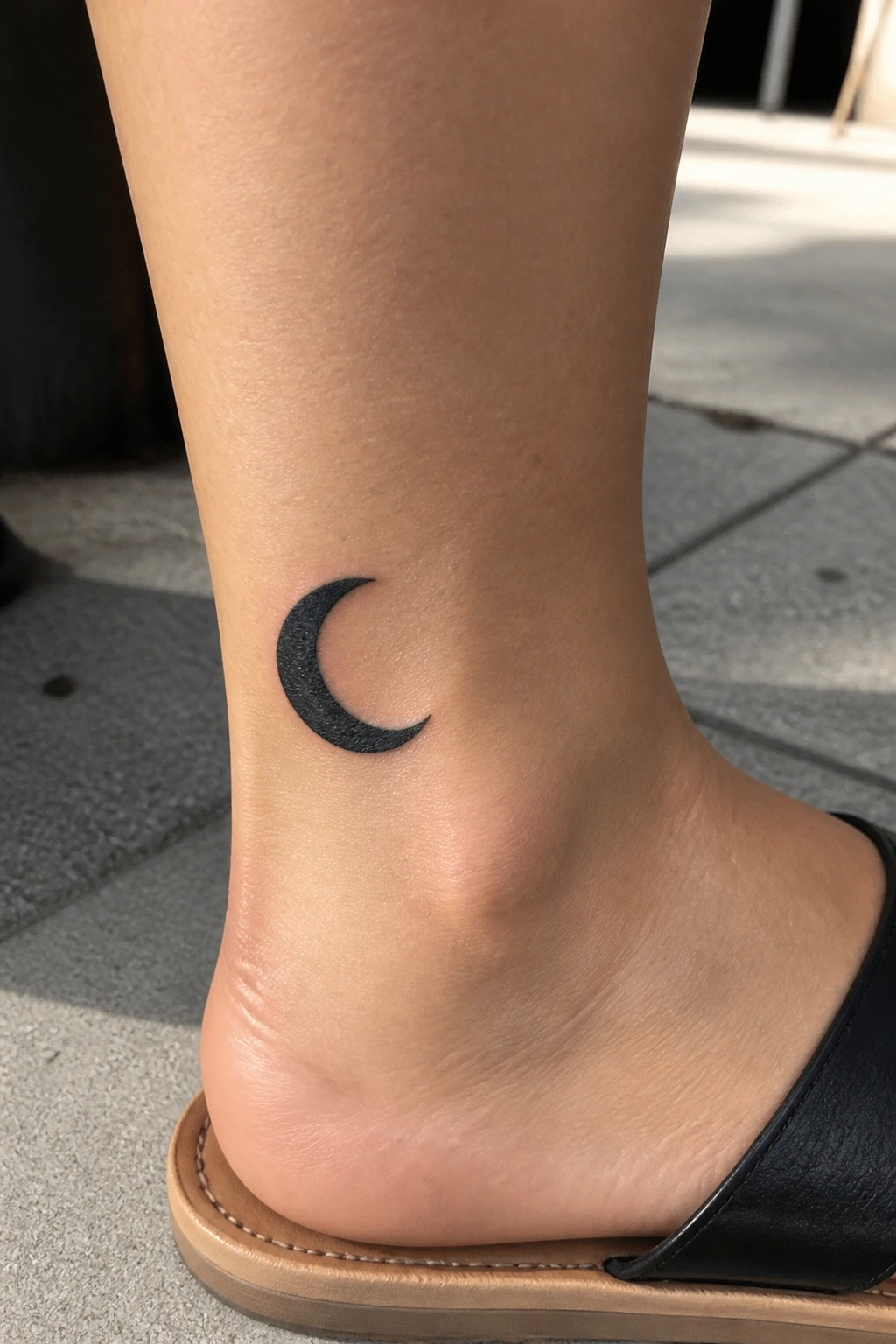 Black crescent moon tattoo on ankle above black slide sandal on foot.