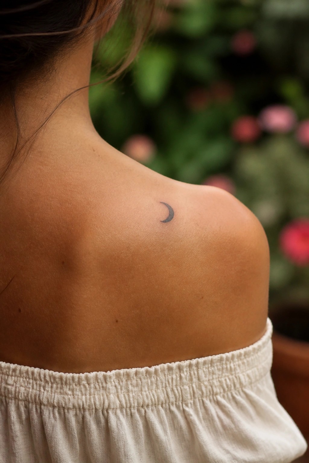 Tanned shoulder with small crescent moon tattoo above white off-shoulder elastic top against blurred blooms.