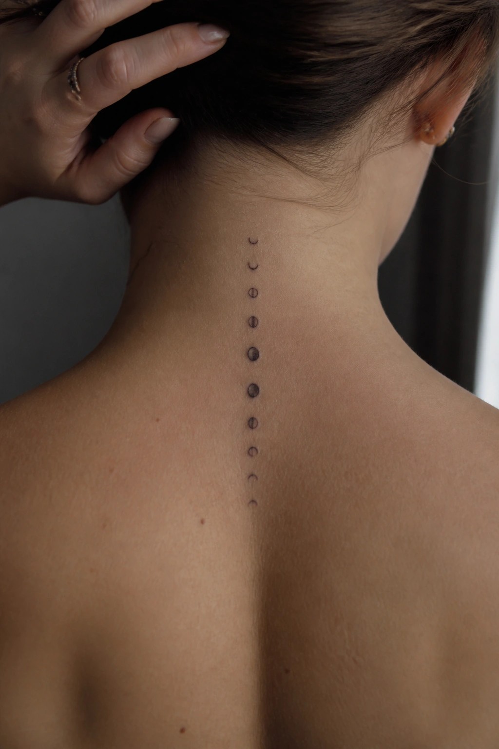 Moon phases tattoo down spine on bare neck and back, hand holding hair
