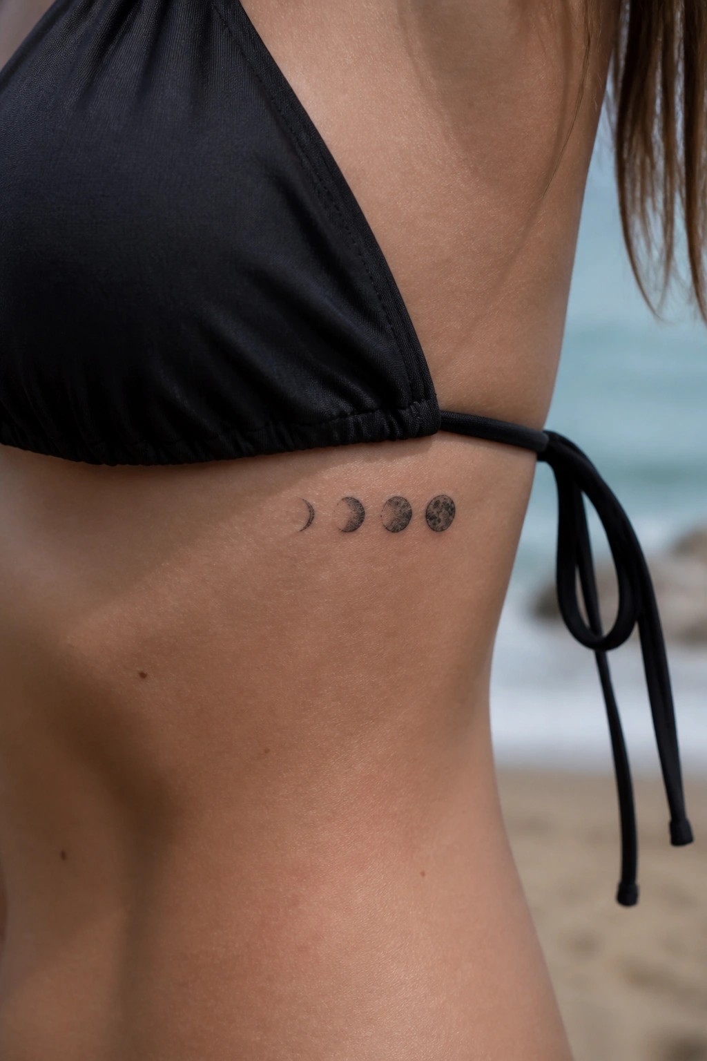 Black bikini top with moon phases tattoo on ribs by ocean