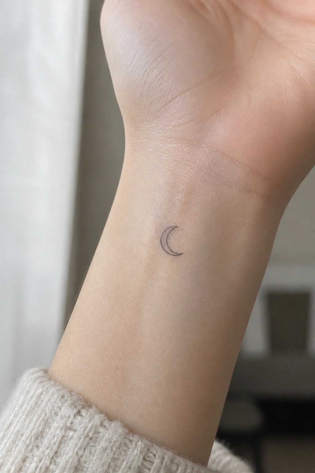 Small crescent moon tattoo on inner wrist above white knitted sweater cuff