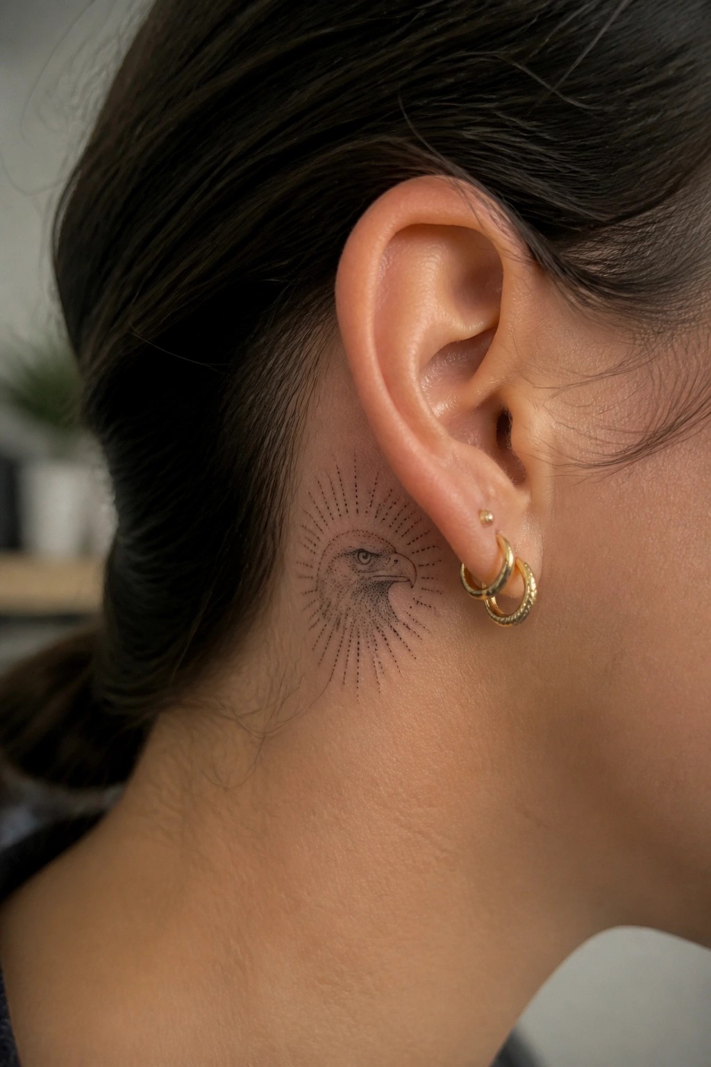 Eagle tattoo with sunburst rays behind ear, gold hoop earrings visible
