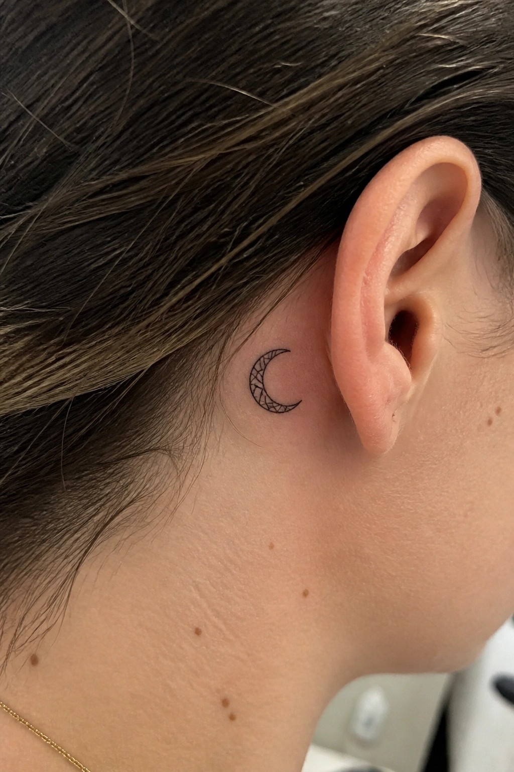 Small crescent moon tattoo with geometric fill placed behind the ear on a woman with dark hair