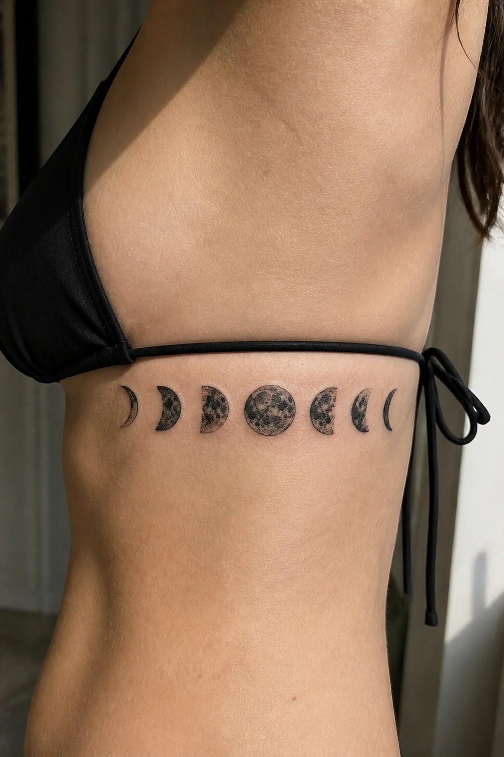 Horizontal row of moon phase tattoos on a woman's side torso above the waistband of black swimwear.