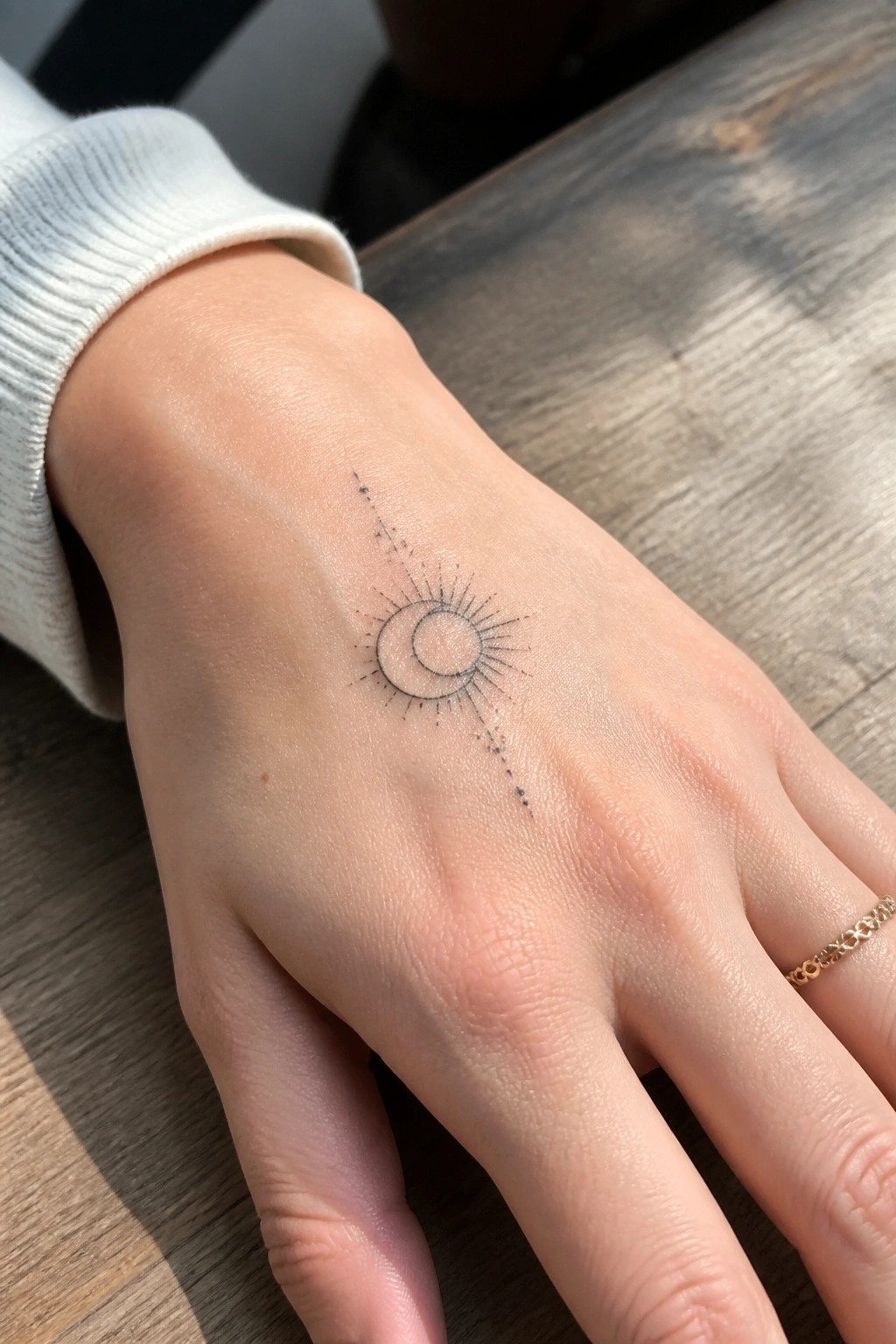 Hand with sun-moon tattoo, white sweater sleeve, gold ring on wooden table.