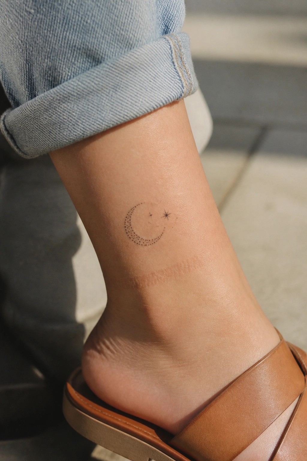 Small dotwork crescent moon tattoo with two stars placed on the inner ankle of a woman wearing rolled jeans and brown sandals.