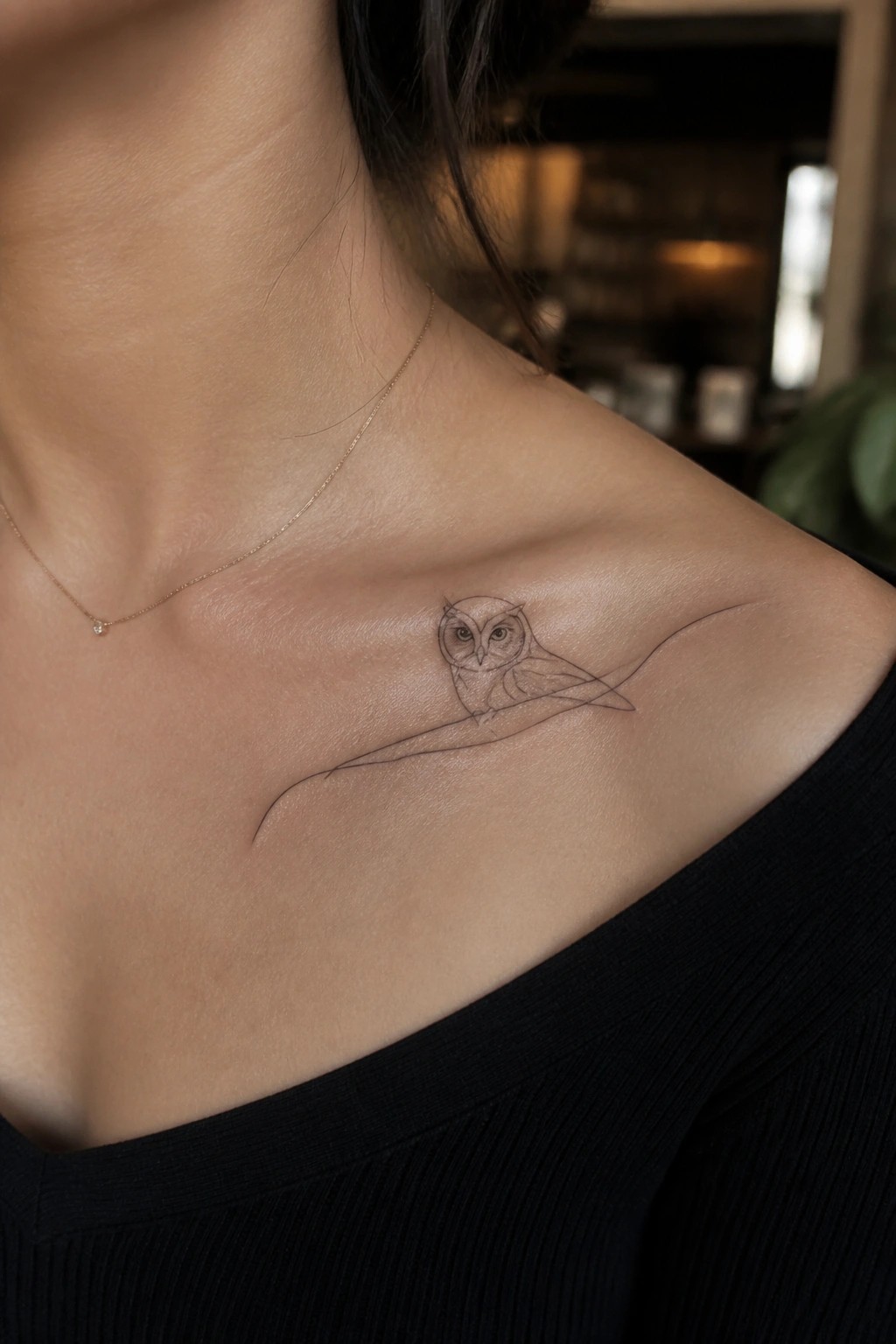 Delicate owl tattoo on woman's collarbone with thin gold necklace and black top