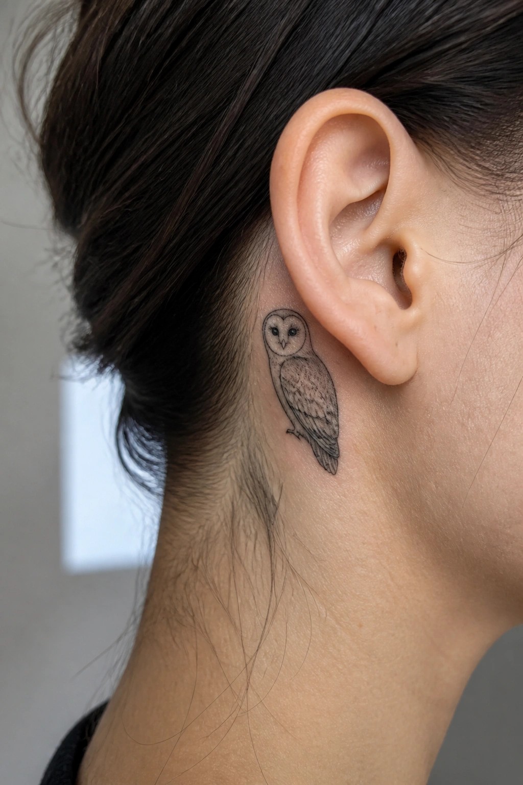 Small black and grey owl tattoo placed behind the ear on the side of the neck.