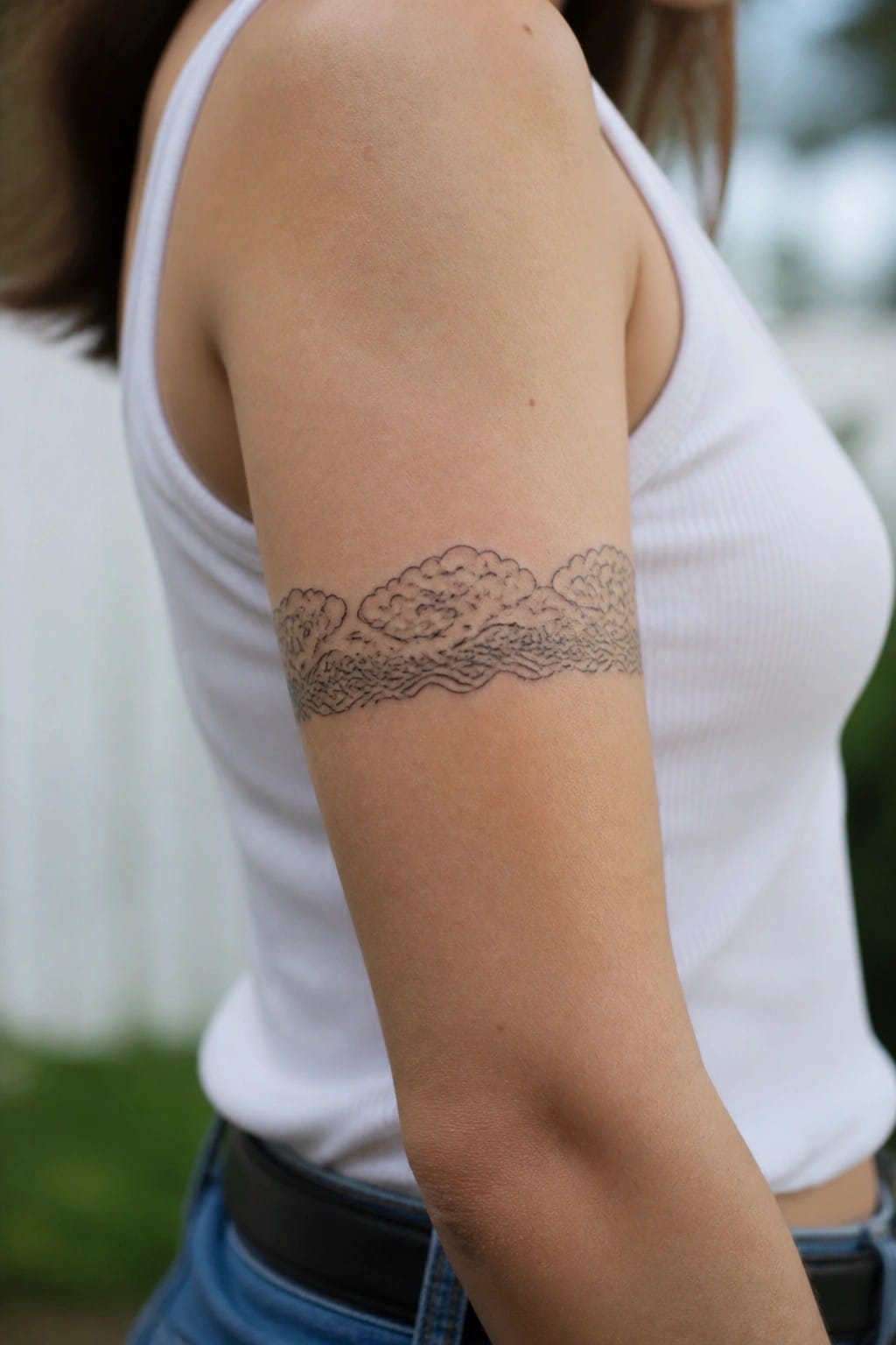Upper arm with black line tattoo of clouds over waves, white tank top.