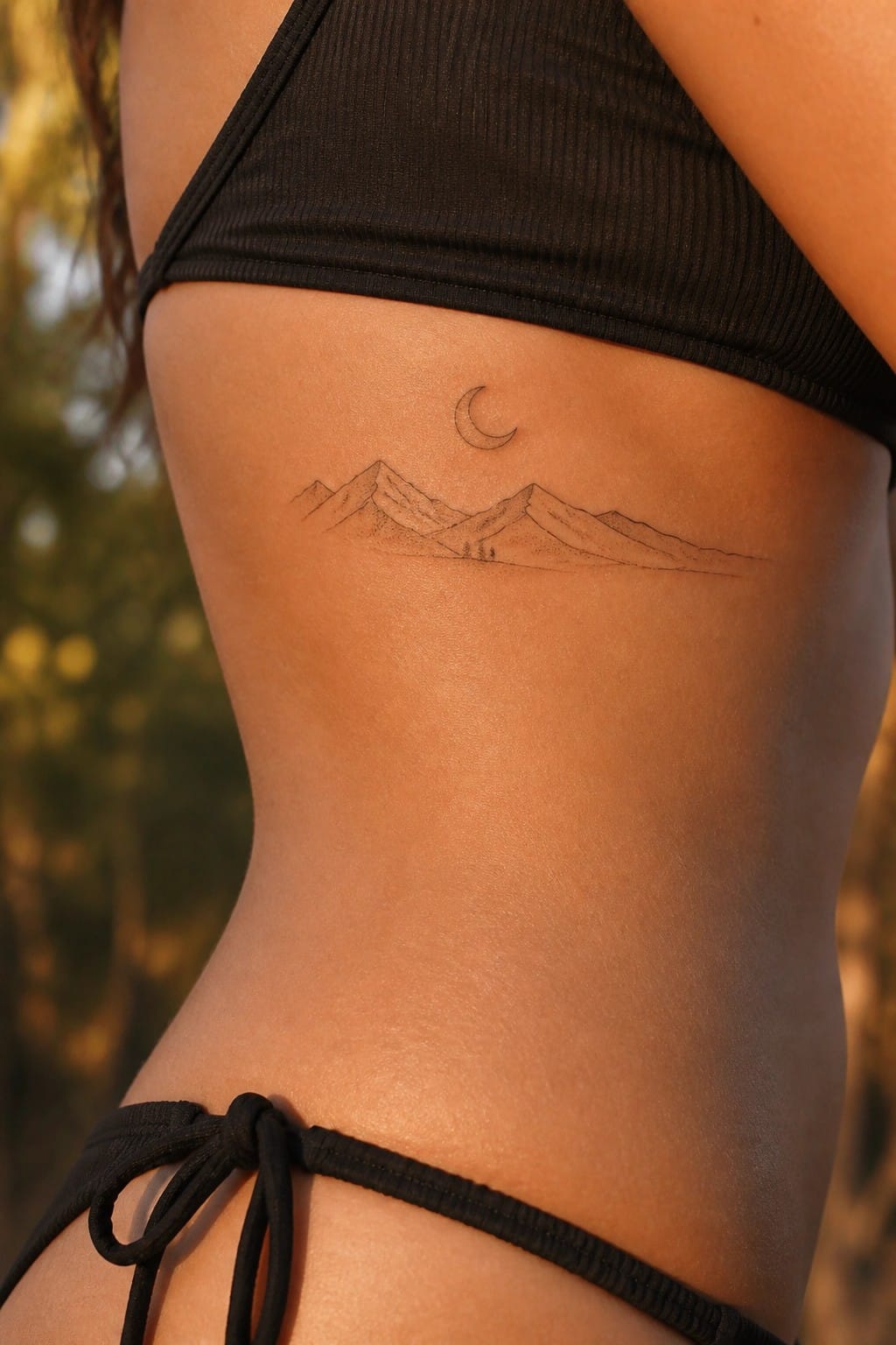 Fine line tattoo of mountains and a crescent moon on the side torso of a woman wearing a black bikini top