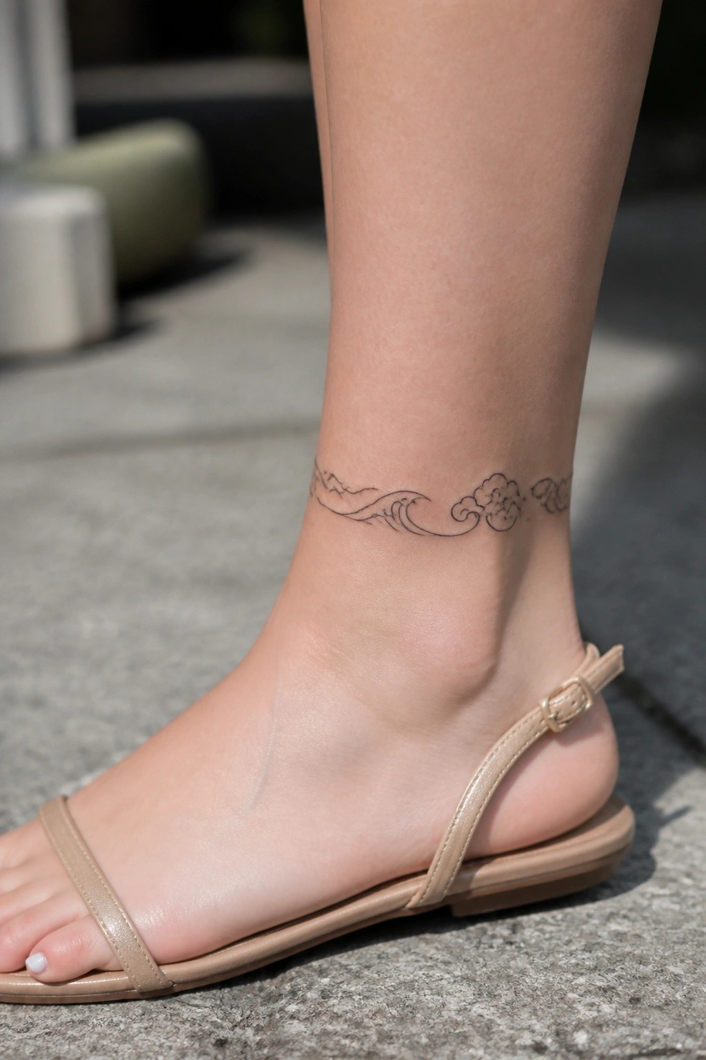 Ankle tattoo of waves and clouds above beige strapped sandal on pavement.