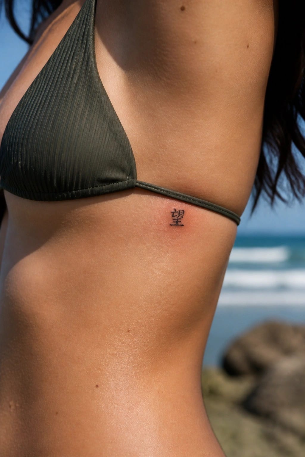 Tanned torso in green bikini with small black Chinese tattoo near ocean waves