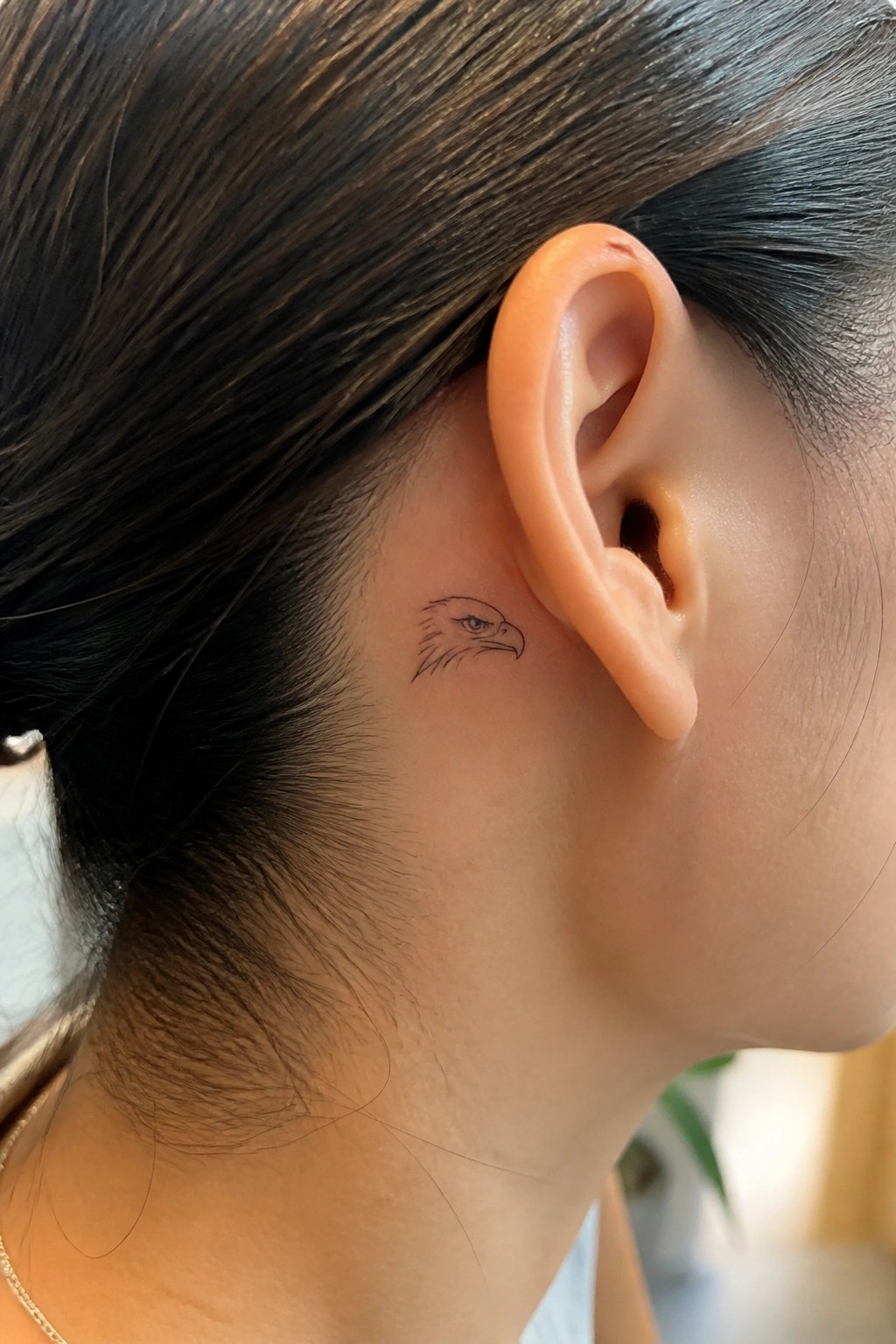 Fine line eagle head tattoo placed behind the ear on a woman with dark hair