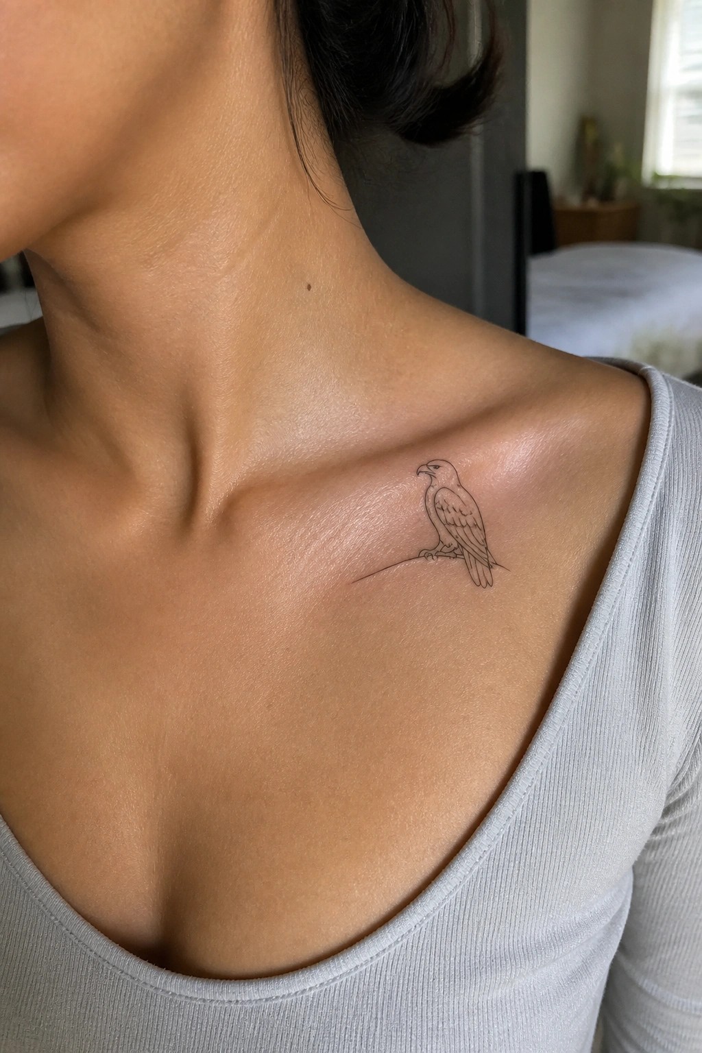 Small eagle tattoo on collarbone above light gray scoop-neck top