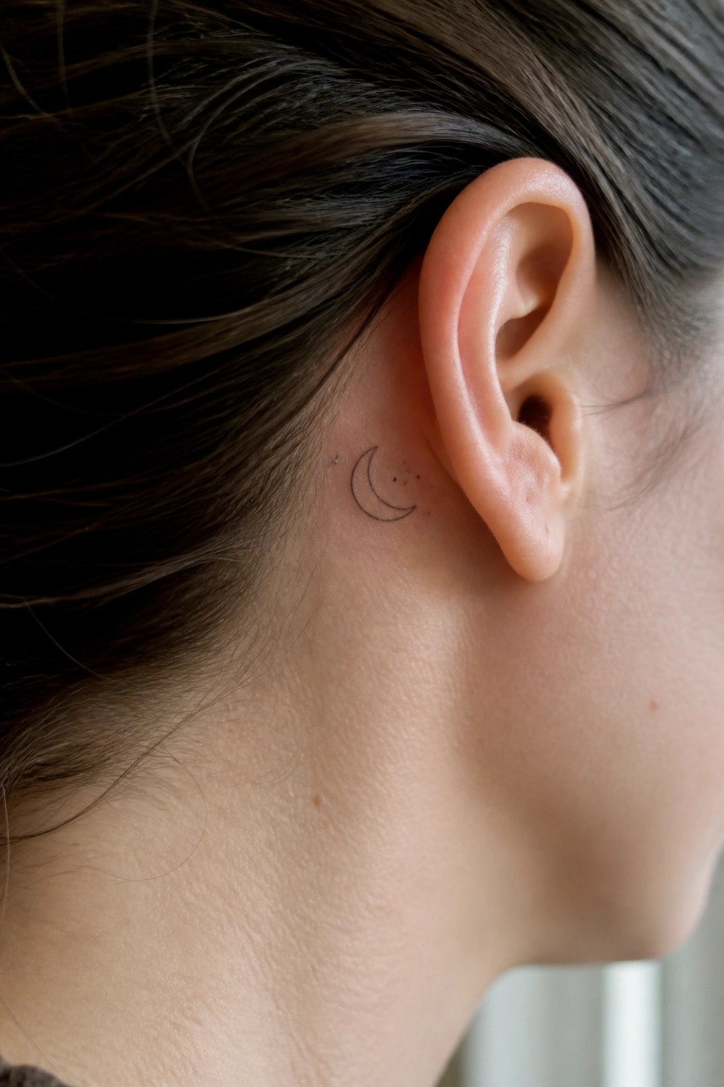 Small fine line crescent moon tattoo with dots placed behind a woman's ear on light skin with dark hair pulled back.