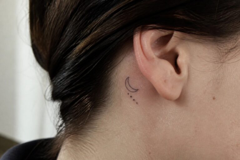 Small fine line crescent moon tattoo with dots placed behind a woman's ear on light skin with dark hair pulled back.