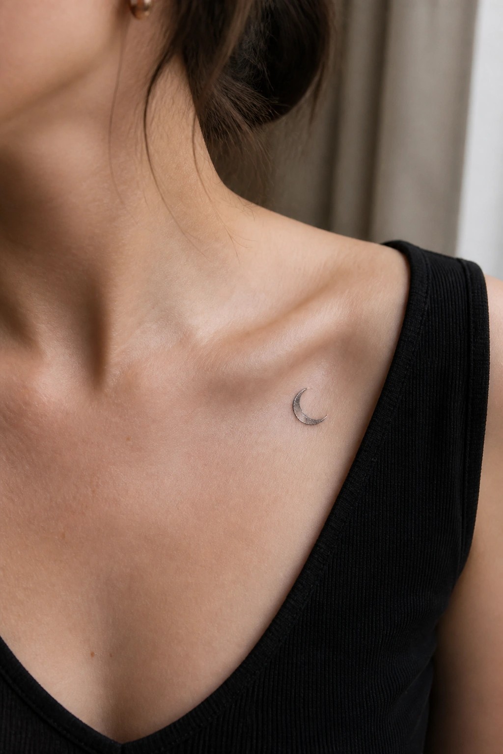 Small crescent moon tattoo on a woman's collarbone area above a black top.
