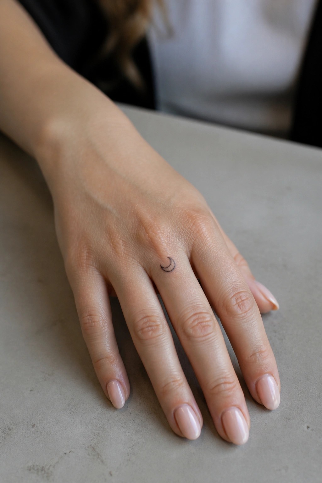Light-skinned hand with crescent moon tattoo on ring finger, pale pink nails on gray surface.