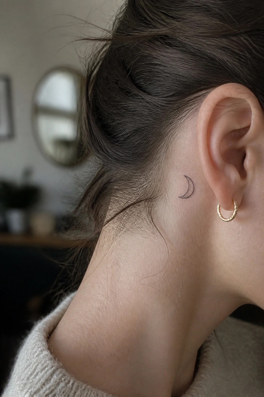 Dark hair pulled back revealing crescent moon tattoo behind ear with gold hoop earring