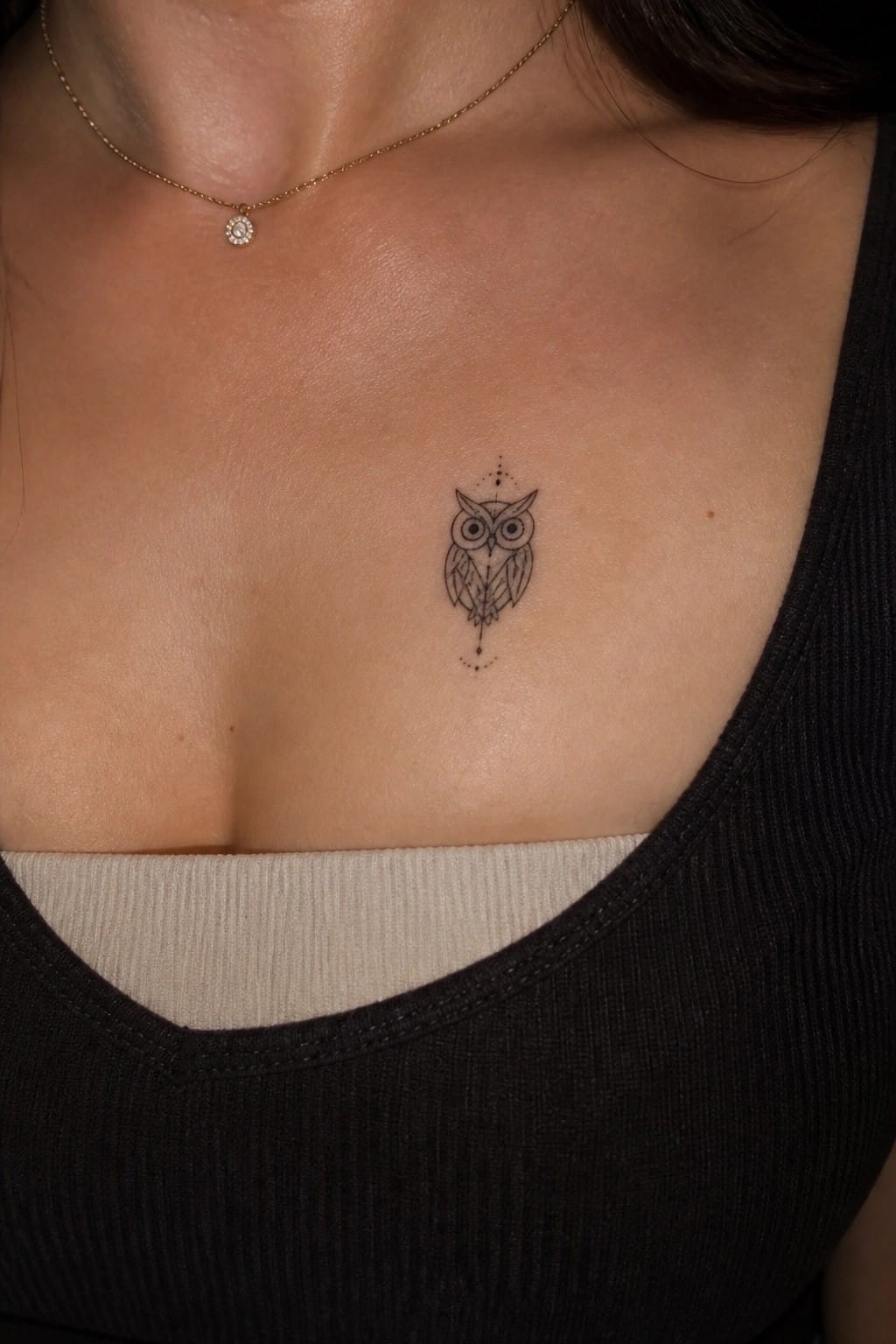 Small black owl tattoo centered on chest above black tank top and gold necklace