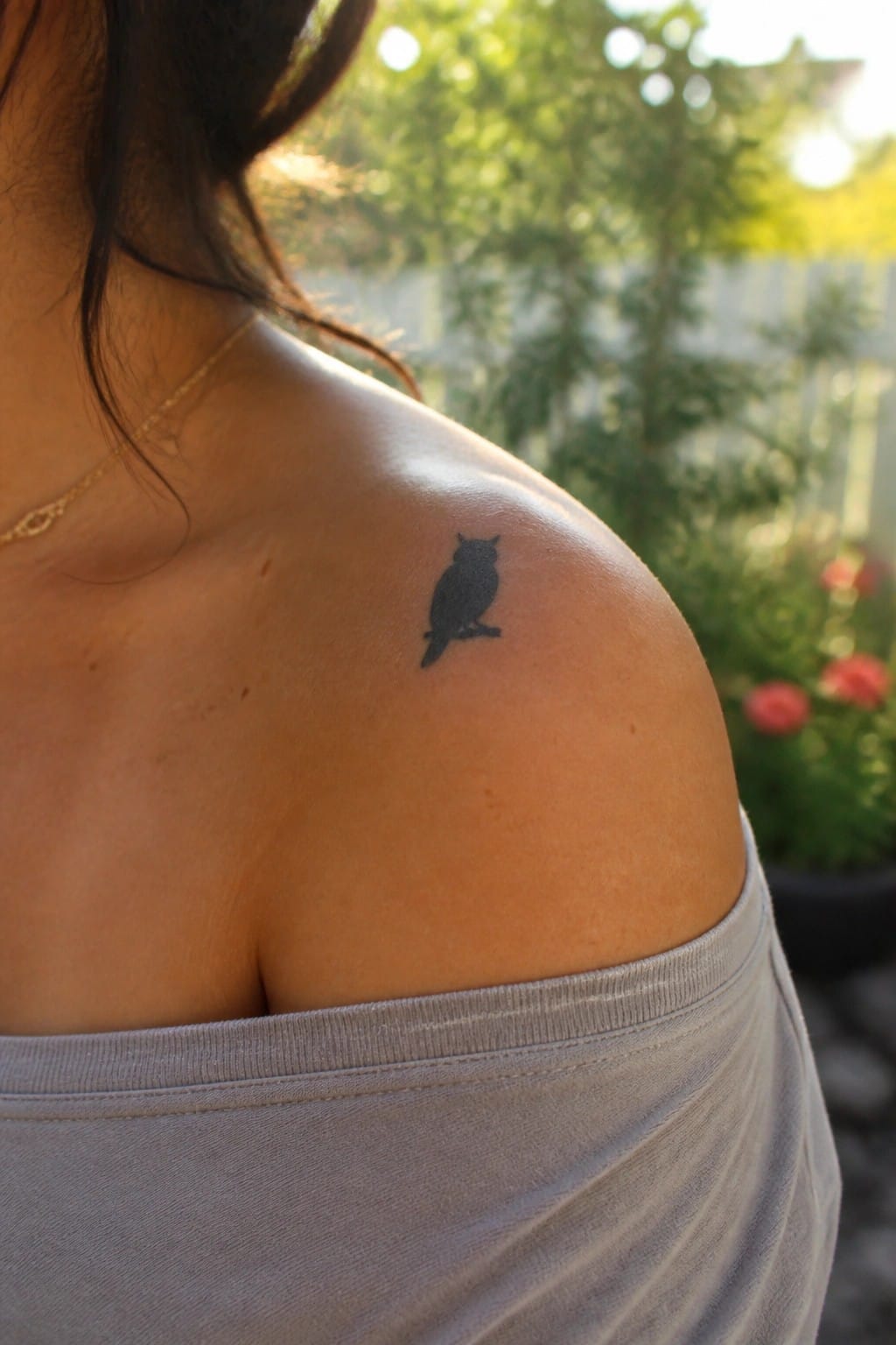 Black owl tattoo on shoulder above gray off-shoulder top with gold necklace
