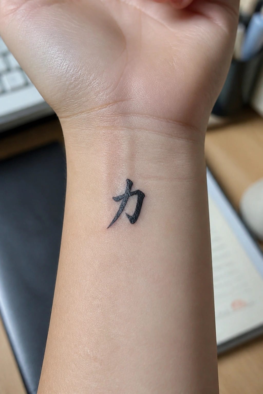 Black Chinese character tattoo on clenched wrist with blurred desk background.